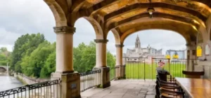 La escena describe una pintoresca plaza con características de arquitectura clásica y elementos naturales en un ambiente sombreado y tranquilo